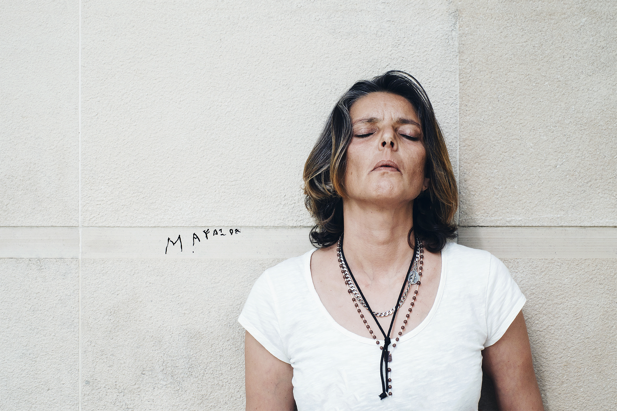 Mulher sentada entre a luz e a sombra encara a câmara, usa três terços ao pescoço.