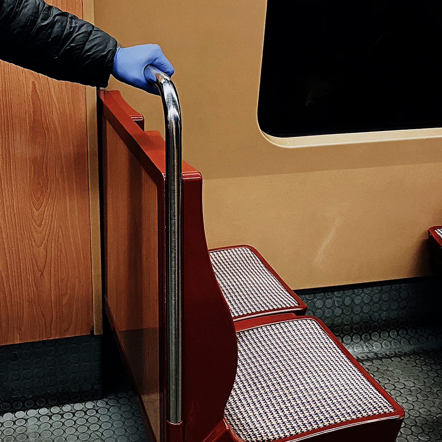 Mão com luva de látex roxa segura em poste horizontal do assento do metro.