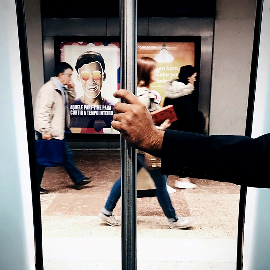 Mão agarra poste vertical do metro, atrás pessoas caminham na plataforma.