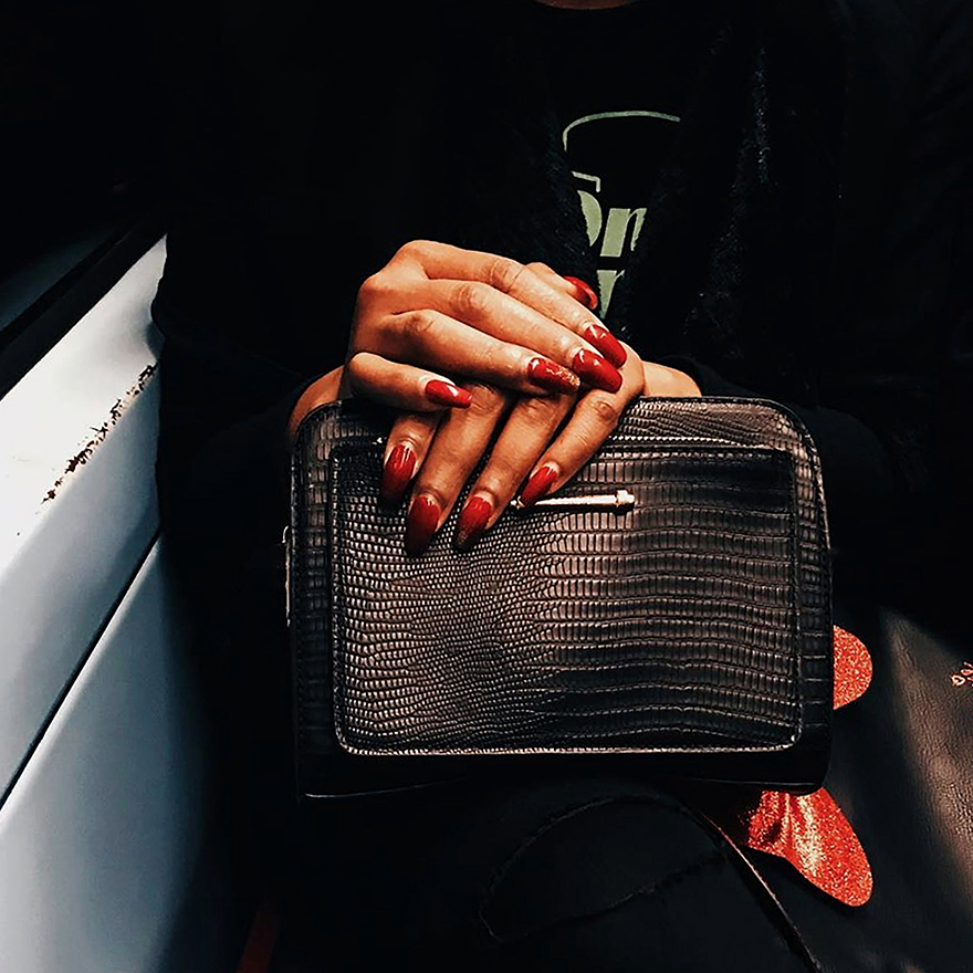 Mãos de mulher com unhas pintadas de vermelho, sobrepostas em cima da sua mala que está no colo.